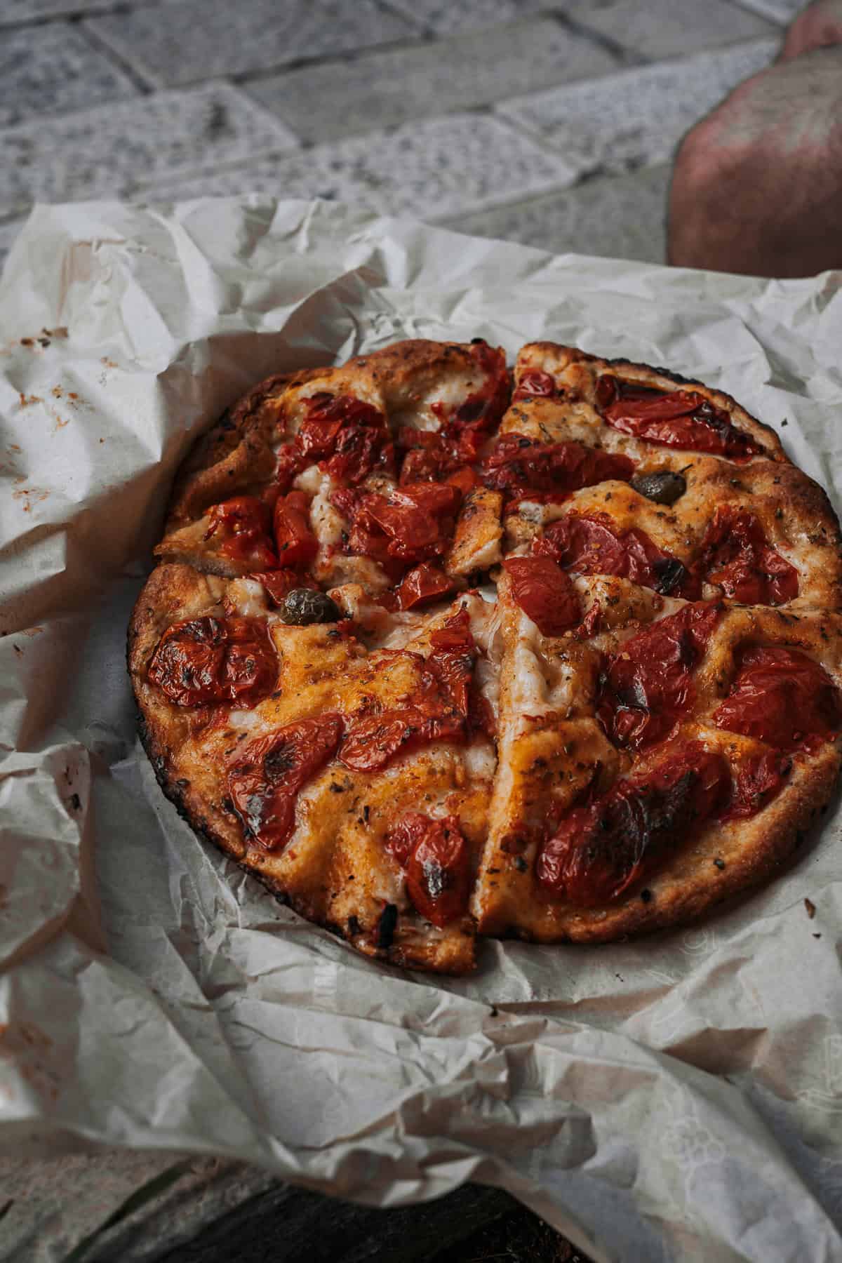 A rustic round focaccia topped with baked cherry tomatoes and green olives is sliced and laid out on parchment, set against stone pavement in an outdoor setting.