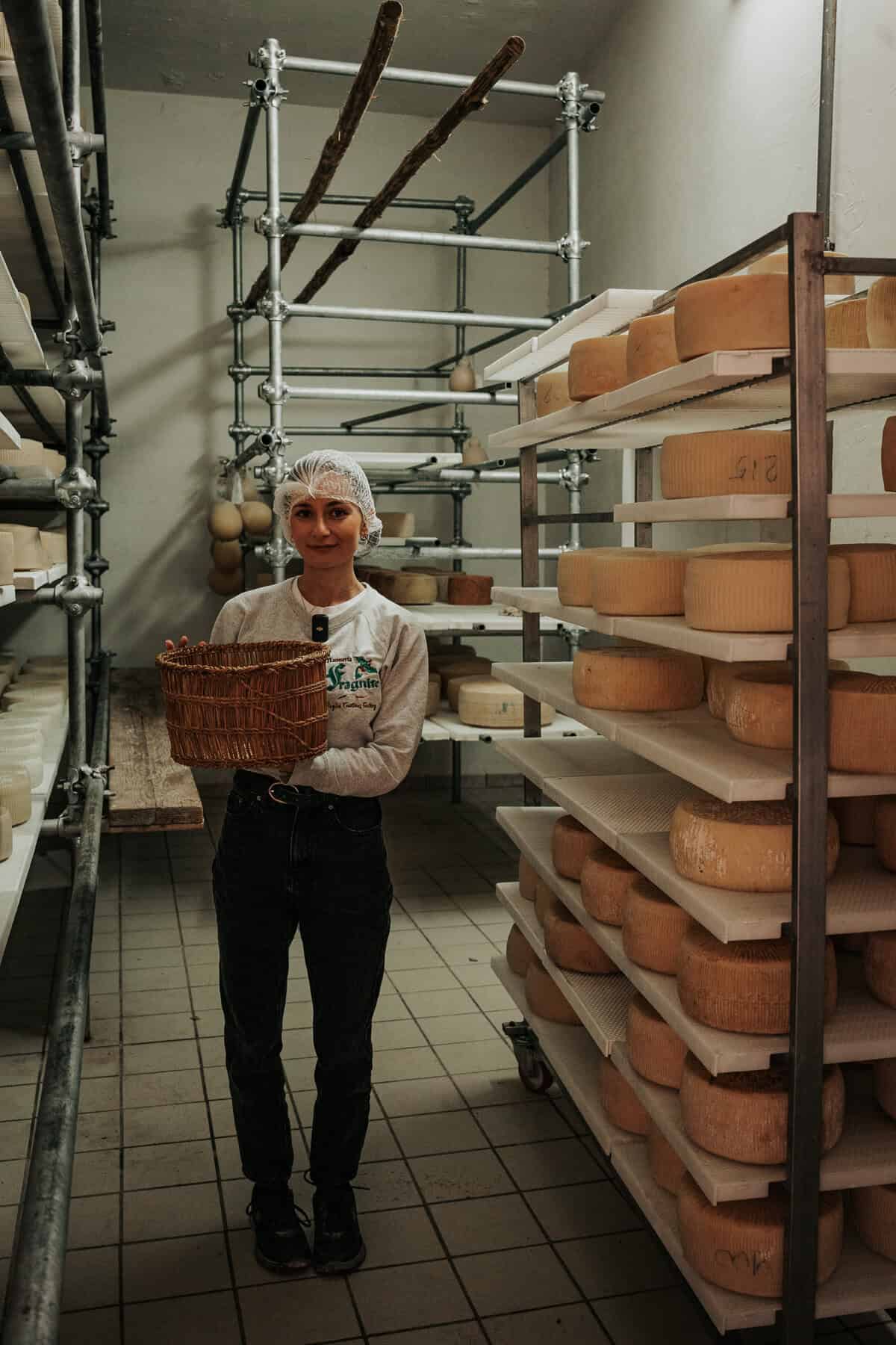 A woman in a cheese aging room holds a wicker basket, surrounded by shelves lined with large wheels of aging cheese on plastic trays.