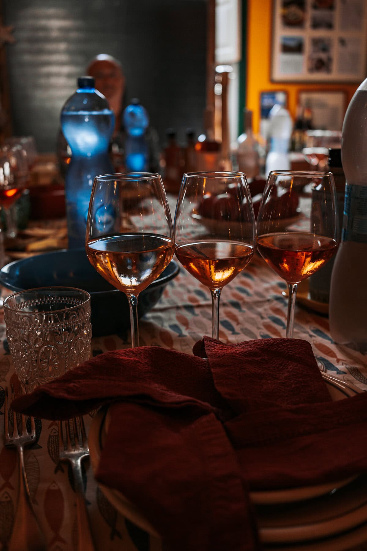 Three glasses of rosé wine sit on a patterned tablecloth surrounded by water bottles, napkins, and cutlery in a cozy, warmly lit dining setting.