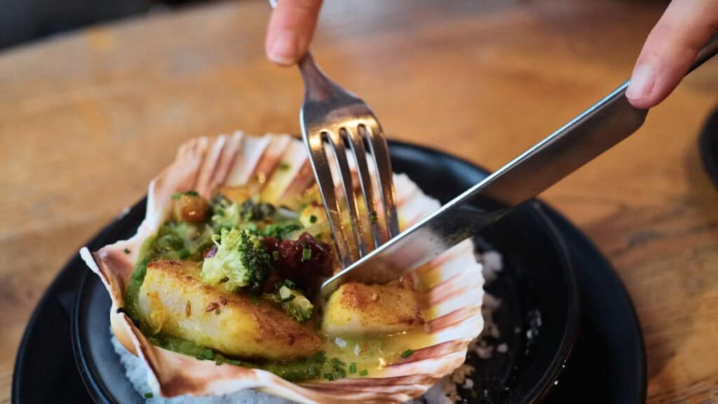 Fork cutting into a perfectly seared Scottish scallop served in its shell with green sauce