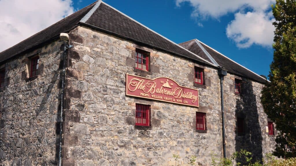 Stone building with red windows and a large sign reading “The Balvenie Distillery Est 1892 Props William Grant & Sons Ltd.” under a bright blue sky. The historic distillery is known for producing Scotch whisky, an important part of traditional Scottish food and drink culture.