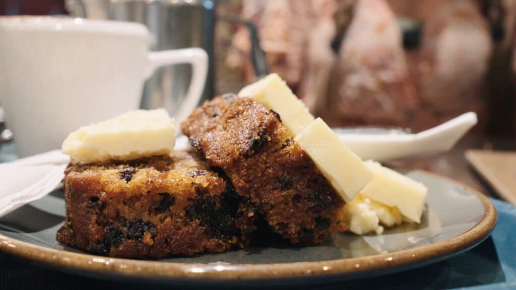 Sticky slices of fruit filled cake topped with thick pieces of butter sit on a plate in a cozy café setting. This traditional English food dessert is served warm with a cup of tea in the background.