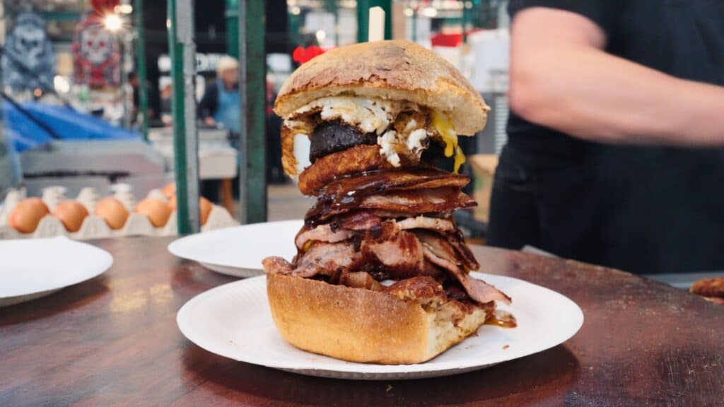 Tall breakfast sandwich stacked with bacon, sausage, black pudding, egg, and potato scone inside a crusty bun on a plate. The towering sandwich showcases a hearty take on traditional Irish breakfast food.