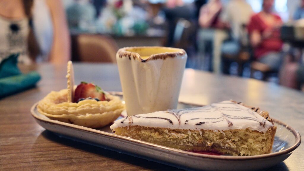 Slice of iced sponge cake served with a small fruit tart and a cup of tea on a café table. This traditional English food dessert is part of a casual afternoon tea setting.