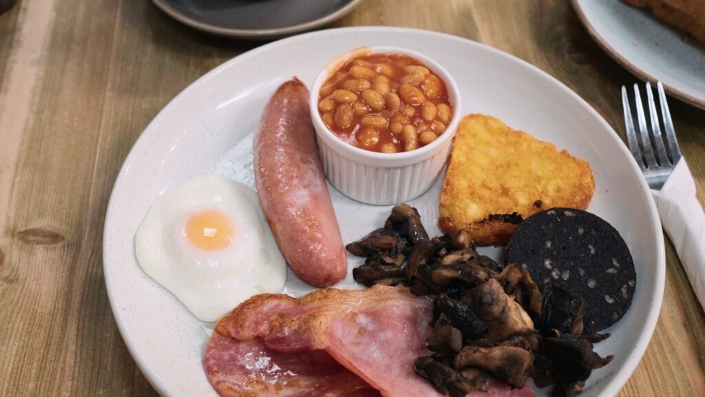 Full English breakfast plate with fried egg sausage baked beans hash brown mushrooms black pudding and bacon arranged on a white plate. This traditional English food meal highlights a classic hearty breakfast.
