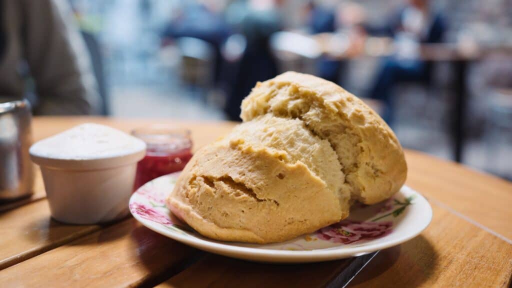 Large golden scone served on a floral plate with clotted cream and red jam on a wooden café table. The freshly baked scone represents a classic Scottish bakery treat often enjoyed with tea.