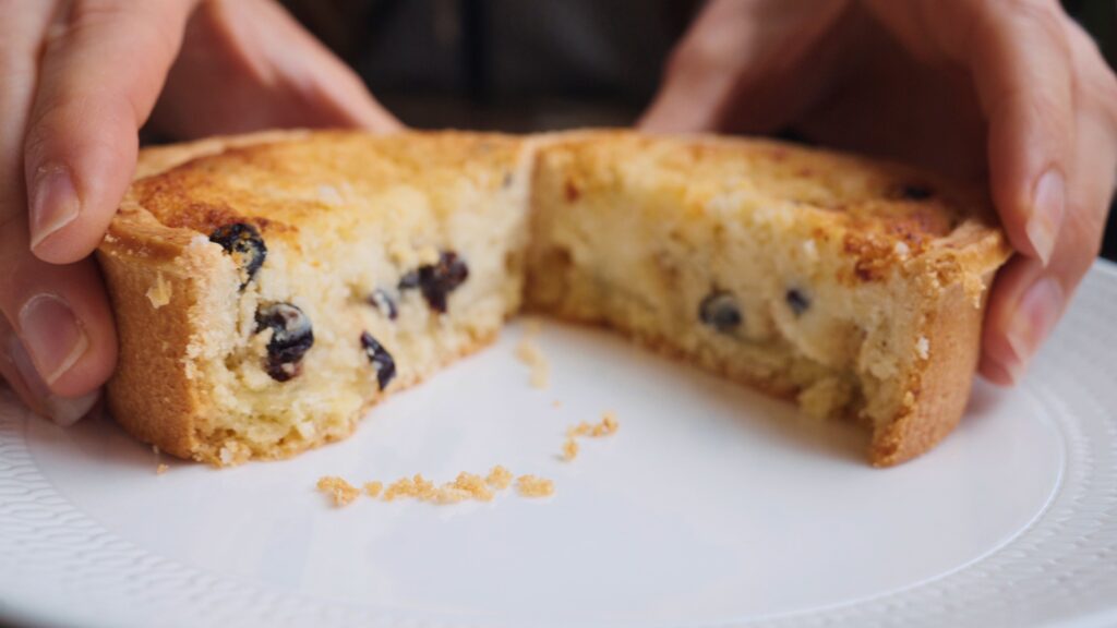 Hands pull apart a small round cake dotted with currants revealing a soft crumb inside on a white plate. This traditional English food bake resembles a currant filled sponge or tea cake.