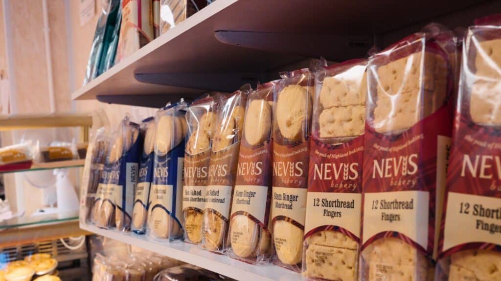 Bakery shelf stocked with packaged shortbread labeled “NEVIS bakery,” “Sea Salted Caramel Shortbread,” “Stem Ginger Shortbread,” and “12 Shortbread Fingers.” Rows of buttery shortbread biscuits showcase a popular Scottish baked treat.