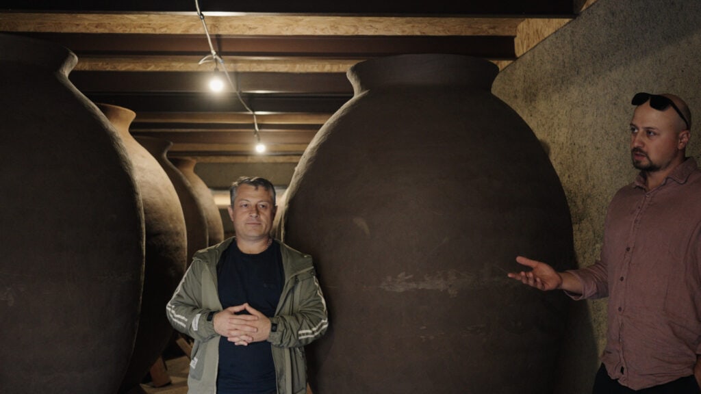 Two men stand inside a dim cellar lined with large clay qvevri used for fermenting wine. This Georgian wine tour moment shows traditional winemaking vessels stored underground.