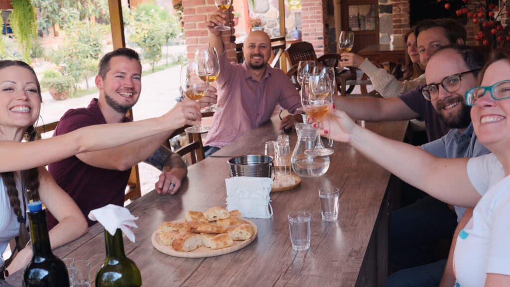 Group of travelers sit around a long wooden table outdoors raising glasses of amber wine in a cheerful toast. This Georgian wine tour captures a shared tasting experience at a vineyard.