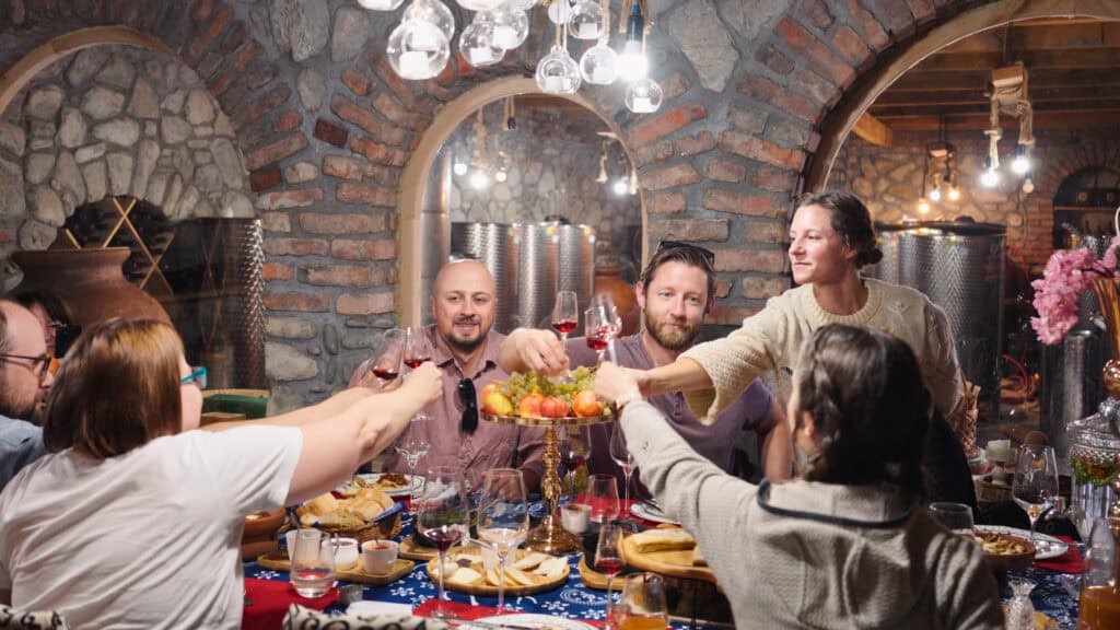 Friends gathered around a rustic table inside a stone cellar clink glasses of red wine over plates of food and fruit. This Georgian wine tour highlights a festive wine tasting in a traditional setting.