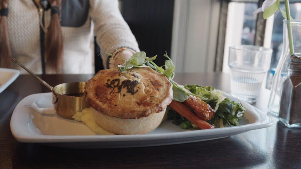 Savory meat pie served with mashed potatoes, roasted carrots, greens, and gravy in a small metal jug. The comforting plate reflects classic pub style traditional Scottish food.