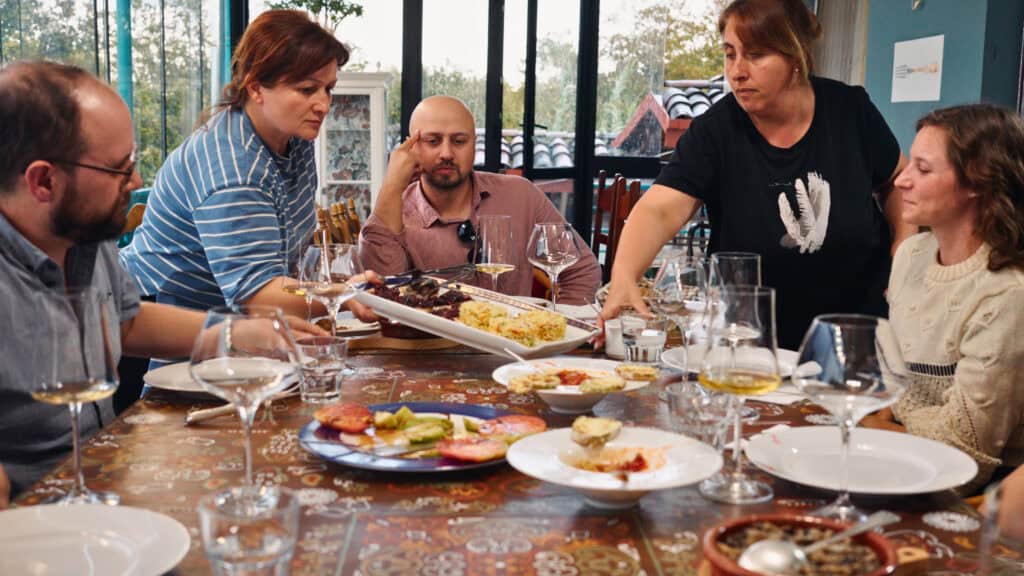 Guests sit around a dining table while a host serves a large platter of food alongside glasses of white wine. This Georgian wine tour scene shows a communal meal paired with local wines.