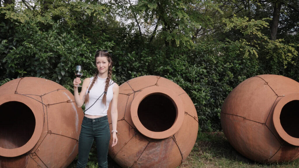 Woman stands outdoors holding a glass of red wine beside large clay qvevri vessels set against greenery. This Georgian wine tour image connects the tasting experience with traditional winemaking methods.