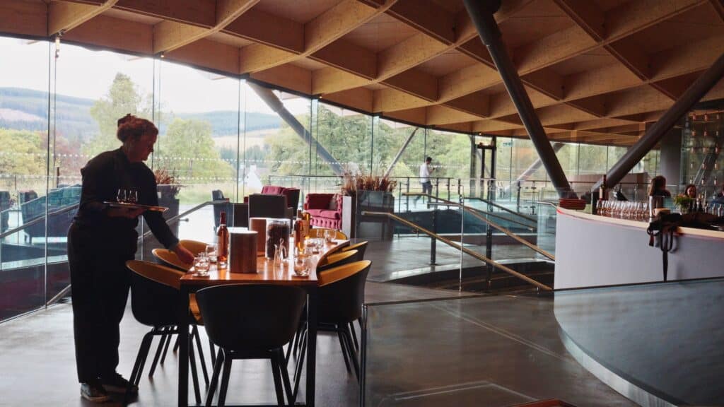 Staff member setting a table with whisky glasses inside a modern tasting room with large glass walls overlooking the Scottish countryside. The tasting experience highlights the connection between Scotch whisky and traditional Scottish food and drink culture.