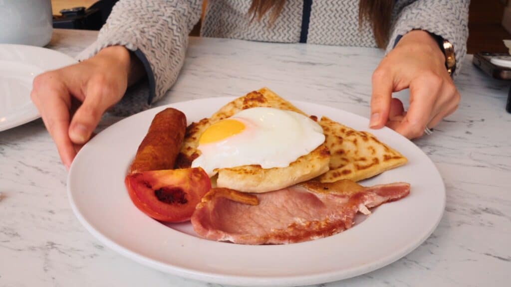Plate with a fried egg, bacon, sausage, grilled tomato, potato scone, and triangular tattie scone on a white plate. The hearty plate is a classic Ulster Fry and a staple of traditional Scottish food.