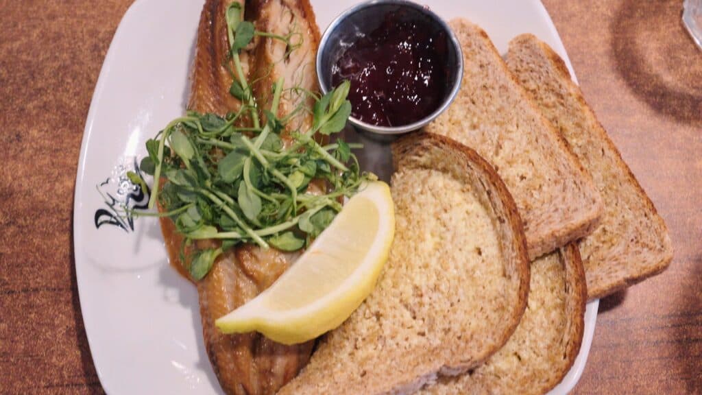Smoked fish fillet garnished with fresh greens and a lemon wedge is served with toasted bread and a small pot of sauce. This traditional English food dish highlights simple ingredients and classic presentation.