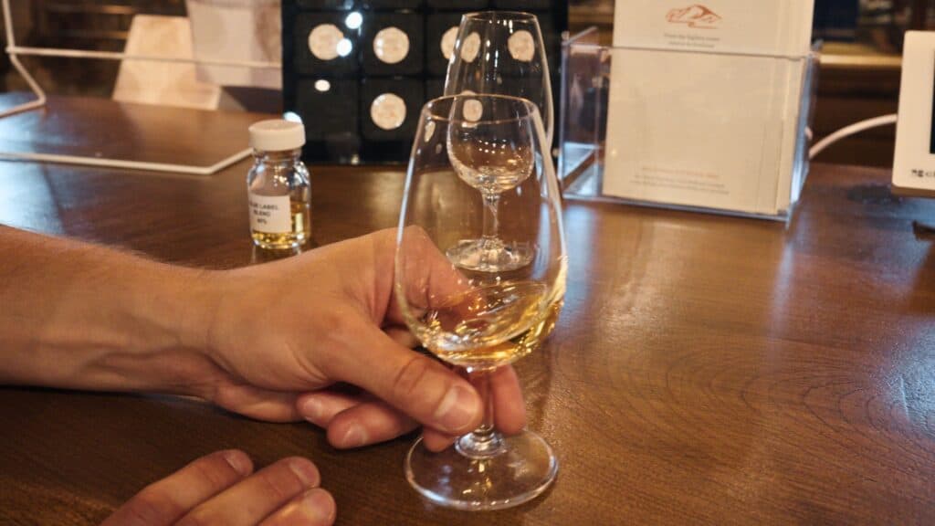 Hand holding a glass of golden whisky at a tasting counter with bottles and display cases behind it. Scotch whisky tasting is closely connected to the culture surrounding traditional Scottish food and