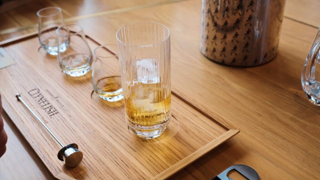 Glass of golden whisky with ice sitting on a wooden tasting tray beside empty whisky glasses and tasting tools. The whisky tasting setup reflects Scotland’s long tradition of whisky as a key part of Scottish food and drink culture.