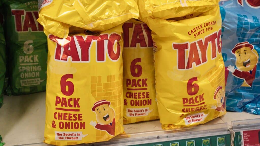 Store shelf stocked with bright yellow bags labeled “Tayto 6 Pack Cheese & Onion” and the slogan “The Secret’s in the Flavour.” The popular crisps are a common snack often enjoyed alongside Scottish comfort foods.