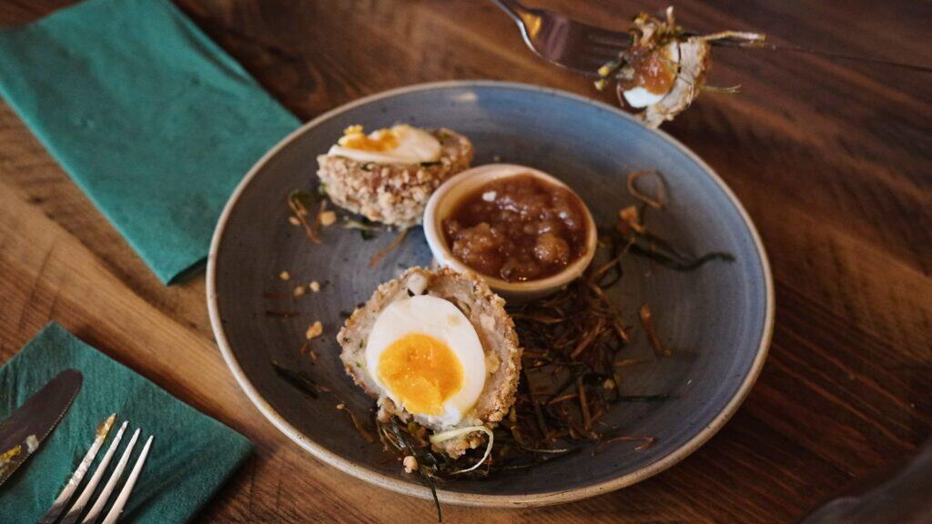 A plated Scotch egg cut in half reveals a soft yolk center beside a small dish of chutney in a restaurant setting. This best restaurants in the Cotswolds dish highlights a refined take on a classic British starter.