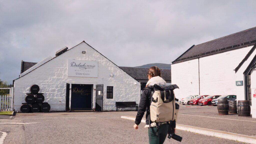 Traveler with a backpack walking toward a white building labeled “Dalwhinnie Distillery Visitor Centre.” Visiting distilleries like Dalwhinnie is a popular way to experience Scottish whisky and traditional Scottish food and drink culture.
