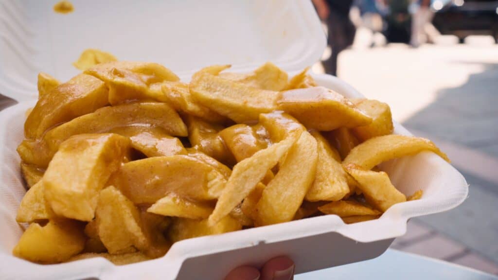 Thick cut chips covered in glossy brown gravy served in a white takeaway container held outdoors. A hearty street style side dish often enjoyed alongside traditional Scottish food.