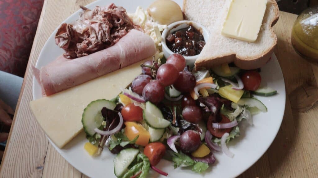 Large plate with sliced ham cheese bread salad and chutney arranged together on a wooden table. This traditional English food spread includes fresh vegetables and simple cold cuts.