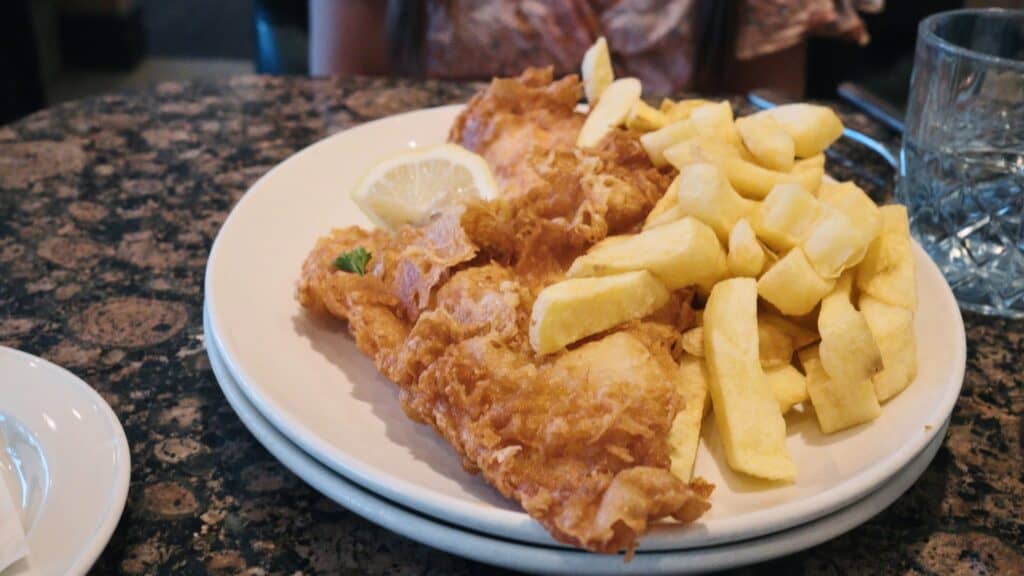 Crispy battered fish served with thick cut chips and a lemon wedge on a white plate. This traditional English food favorite is a classic fish and chips meal.