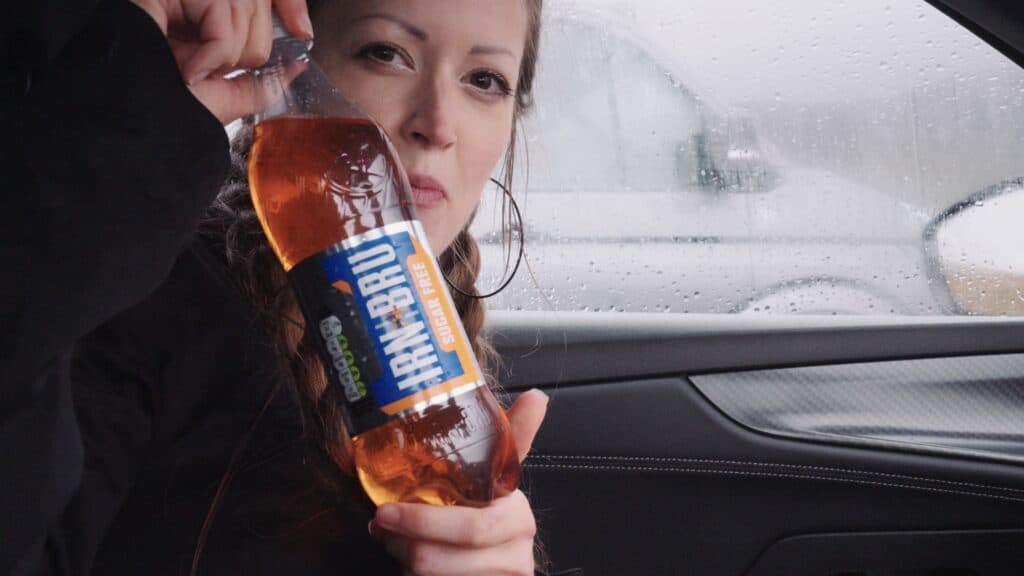Person sitting in a car holding a bottle labeled “IRN BRU Sugar Free.” The bright orange soft drink is a well known Scottish soda commonly paired with traditional Scottish food.