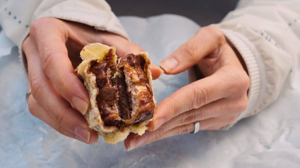 Hands breaking open a deep fried chocolate bar to reveal melted chocolate and caramel inside the batter. The indulgent treat is a famous Scottish takeaway dessert often associated with traditional Scottish food culture.