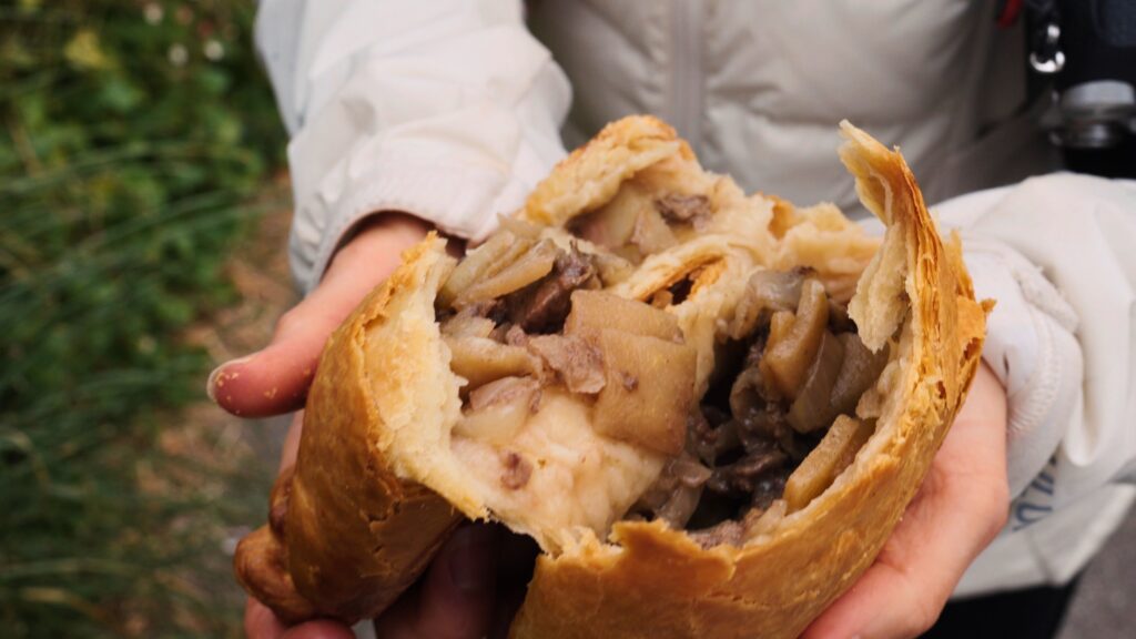 Hands hold a freshly baked pastry broken open to reveal chunks of meat and potatoes inside a flaky crust. This traditional English food pasty is shown outdoors highlighting its hearty filling.