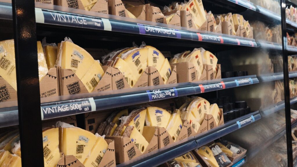 helves in a shop display large wedges of cheddar cheese in branded packaging with labels reading "Vintage" and "Cave Matured 500g". This traditional English food selection showcases different aged cheeses available for purchase.