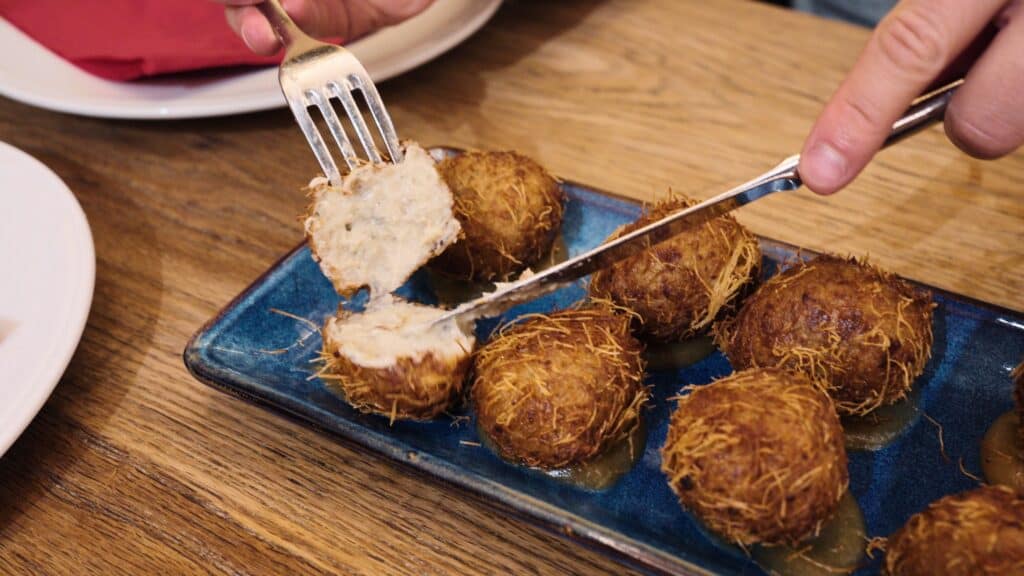 Crispy croquettes are cut open to reveal a creamy filling, arranged on a blue plate on a wooden table. This classic Spanish dish is a must try at restaurants in calle ibiza madrid.