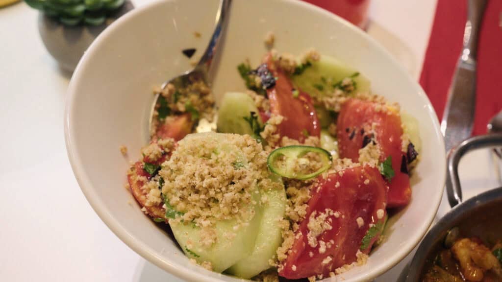 : Fresh tomato and cucumber salad topped with herbs and breadcrumbs served in a white bowl, a simple dish representing traditional Georgian foods and fresh local ingredients.