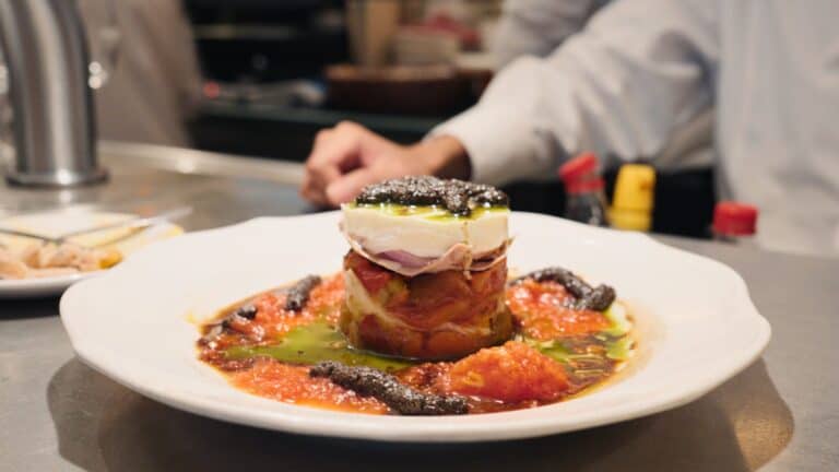 A plated tapa with layers of tomato, fish, and a soft cheese topped with dark caviar and olive oil sits on a white dish at the bar. This refined presentation highlights the creative small plates found in restaurants in calle ibiza madrid.