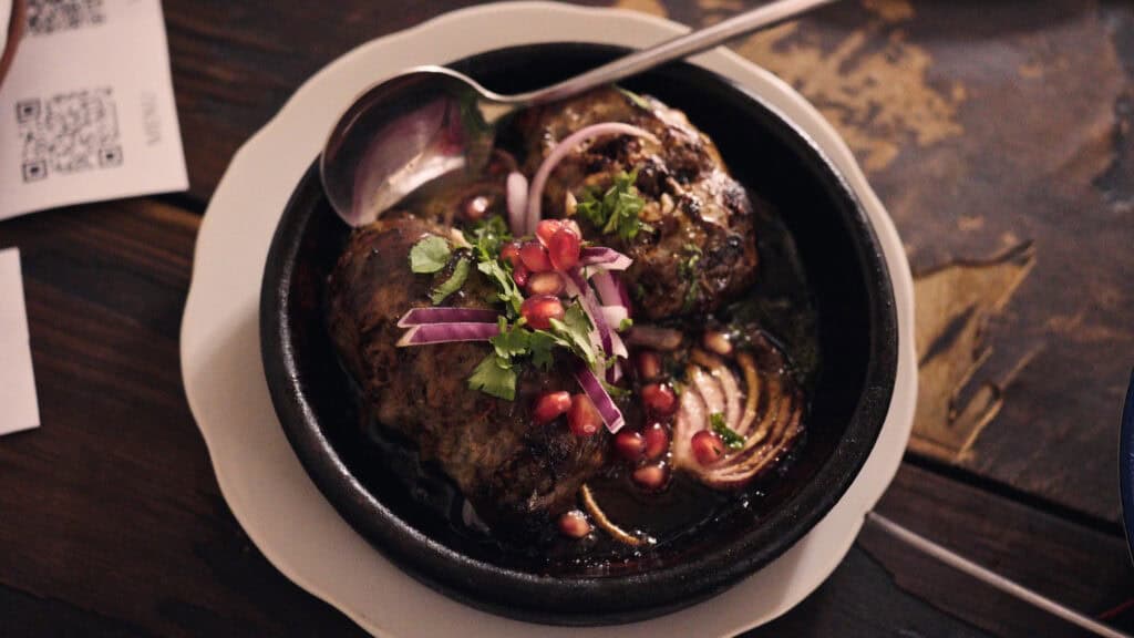Clay dish with roasted meat topped with sliced onions, herbs, and pomegranate seeds in a rich sauce, a flavorful example of traditional Georgian foods.