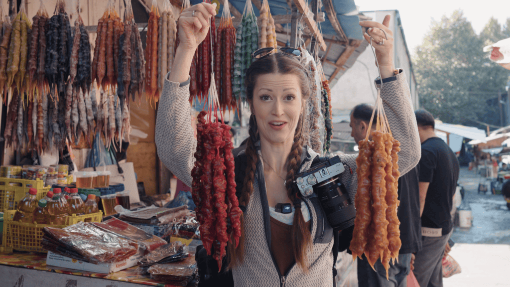 Woman smiling at an outdoor market holding two strings of colorful candied nuts with more hanging behind her, showcasing a popular sweet from traditional Georgian foods.