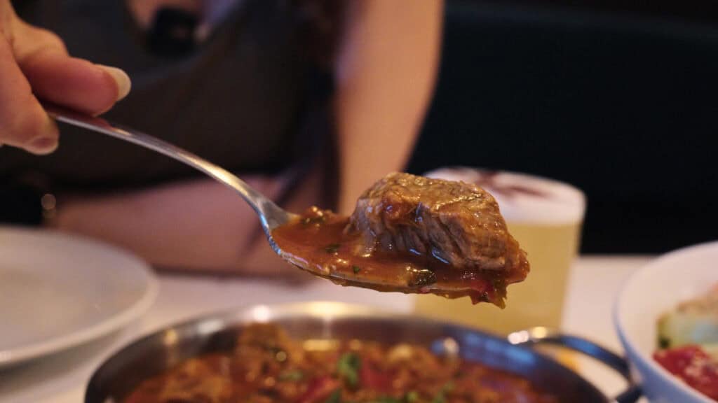 Spoon holding a tender chunk of beef in a thick savory sauce above a pan, showcasing slow cooked meat typical of traditional Georgian foods.