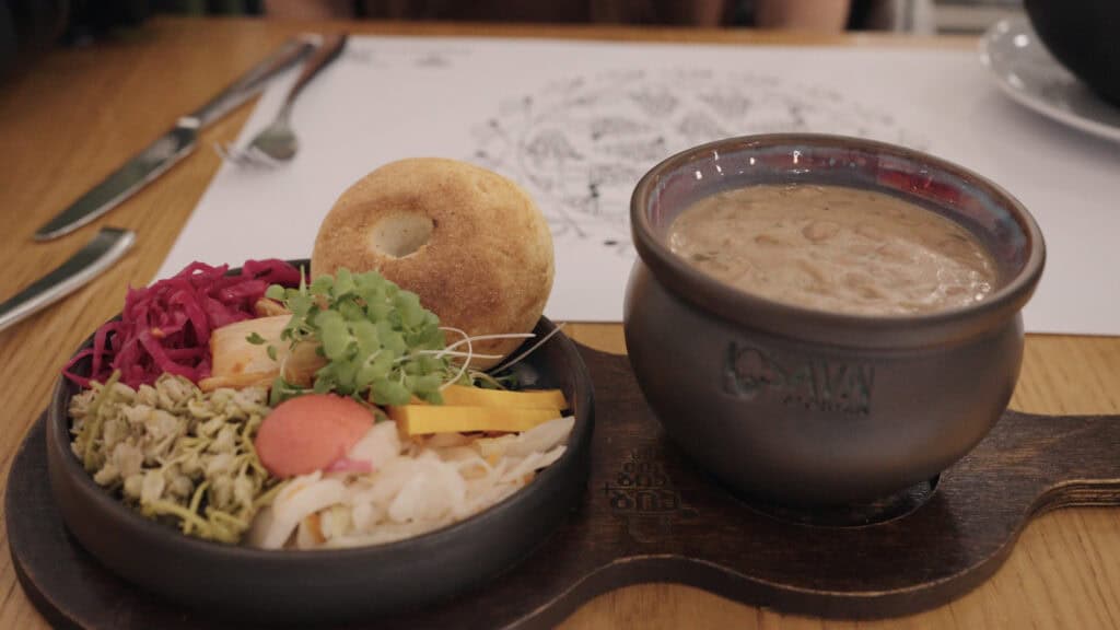 Wooden serving board with a bowl of creamy bean stew, pickled vegetables, fresh herbs, and a small bread roll, illustrating comforting traditional Georgian foods.