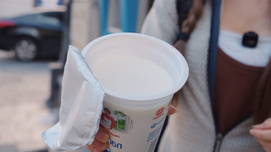 Hand holding a plastic cup of a white yogurt like drink with Georgian writing on the label, a common beverage alongside traditional Georgian foods.