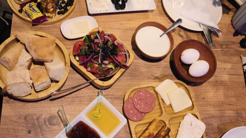 Wooden table spread with bread, cheese, eggs, cured meats, honey, and a tomato salad, showcasing a variety of traditional Georgian foods served family style.