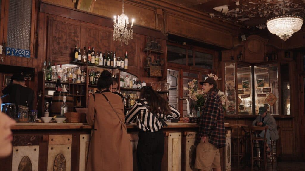 Warm wood lined cocktail bar in Barcelona with chandeliers hanging overhead and people gathered around the counter ordering drinks. Cozy historic interiors like this are part of where to eat in Barcelona for late night drinks and tapas.