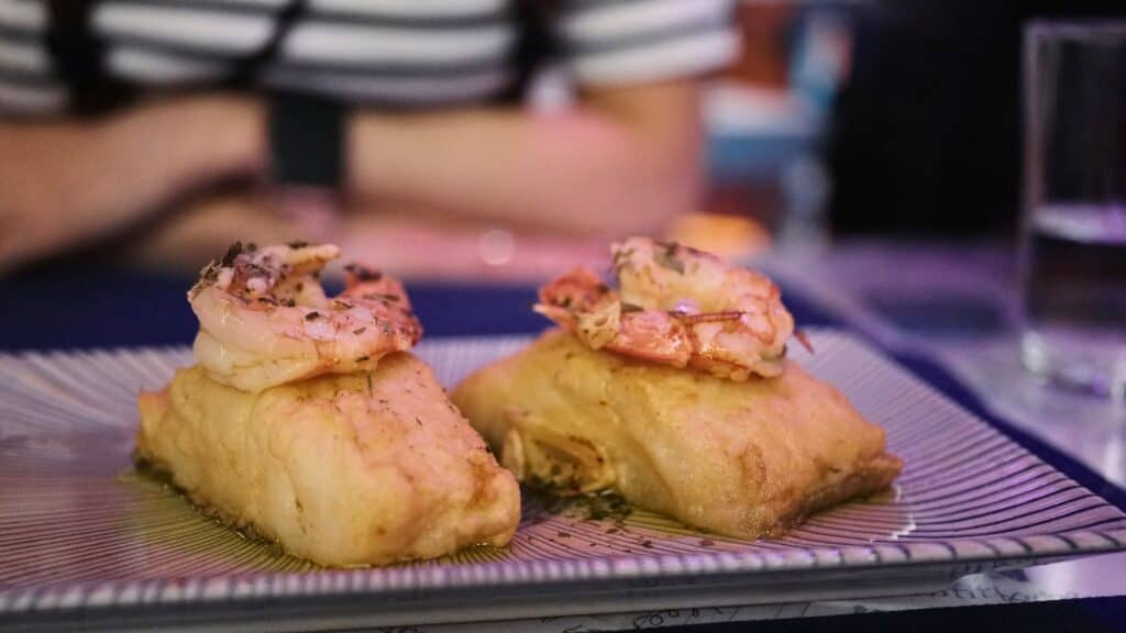 Two pieces of soft bread topped with grilled shrimp and herbs served on a striped plate under dim restaurant lighting. Seafood tapas like these can make the cost of food in Madrid higher at trendy restaurants.