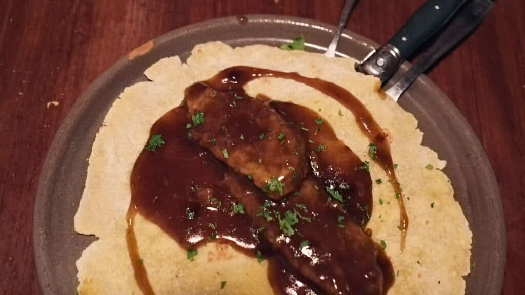 Two pieces of meat covered in glossy brown gravy and sprinkled with herbs on top of a crisp flatbread. The rich sauce and rustic plating showcase hearty dishes served at some of the best restaurants in Bilbao.