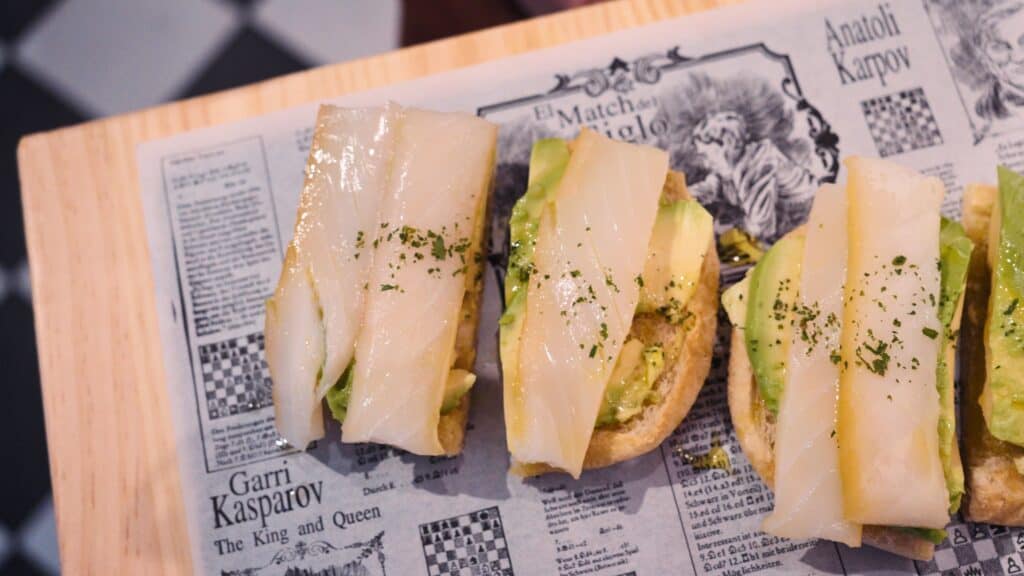 Three small toasts topped with avocado and thin slices of white fish are arranged on paper printed with chess themed text including "Anatolij Karpov" and "Garri Kasparov The King and Queen." The artistic presentation reflects the modern tapas style found in restaurants in calle ibiza madrid.