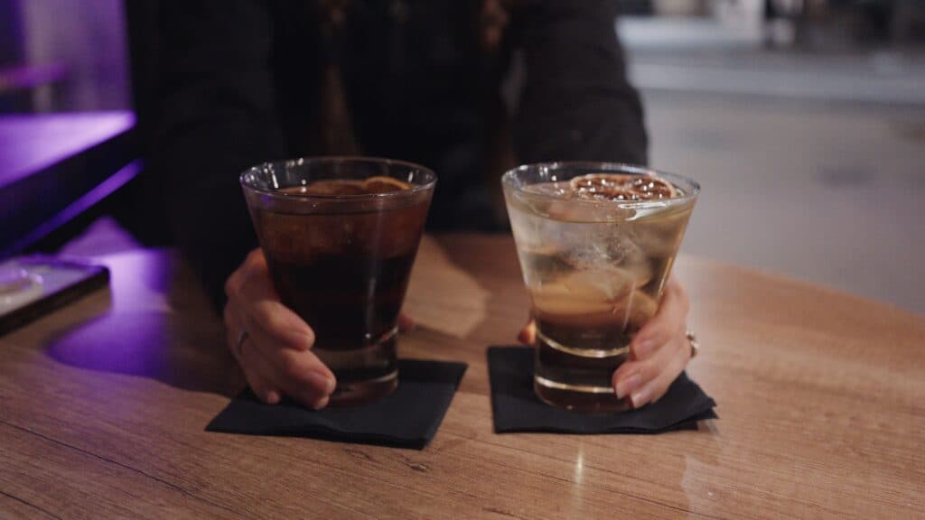 Two cocktails served on a wooden table with large ice cubes and citrus garnish in a dimly lit bar. Classic cocktails and intimate bars are part of the best restaurants in Barcelona nightlife scene.