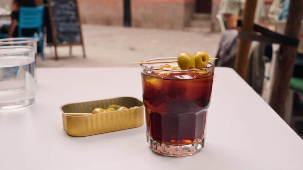 Glass of vermouth with olives served over ice at an outdoor table beside a small tin of olives. Classic aperitif drinks like this are a popular and affordable part of the cost of food in Madrid.