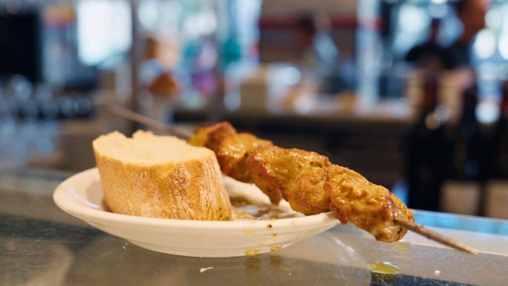 Skewer of grilled marinated meat served with thick sliced bread on a small white plate at a busy bar counter. The warm lighting and casual presentation capture the pintxo culture found in the best restaurants in Bilbao.