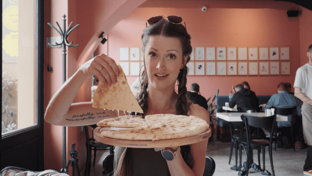 Woman in a restaurant lifting a slice of cheesy bread from a round pie on a wooden board, melted cheese stretching as she serves a classic dish from traditional Georgian foods.